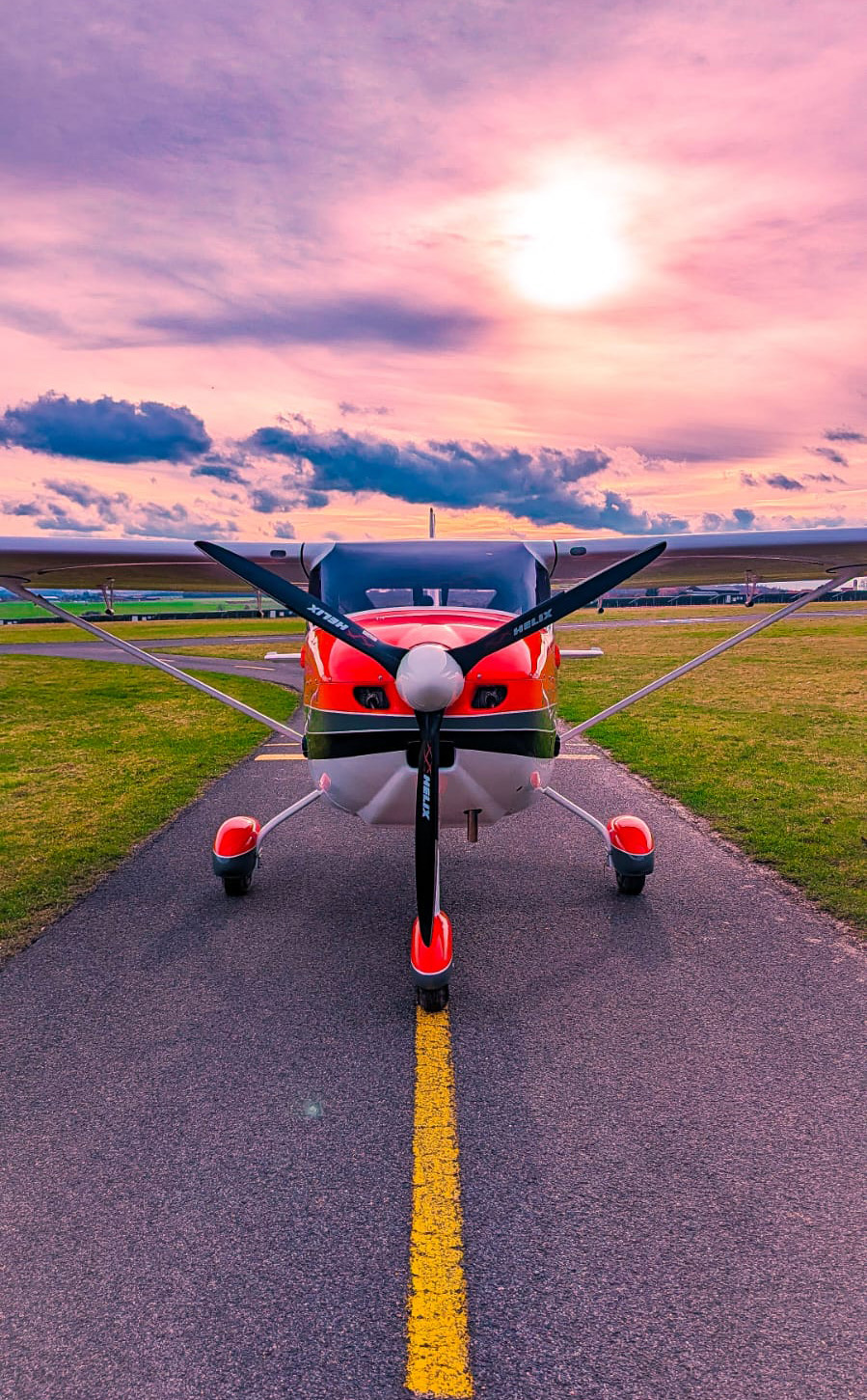 Frontansicht eines roten Leichtflugzeugs auf dem Rollweg bei Sonnenuntergang, mit dramatisch rosa gefärbtem Himmel im Hintergrund.