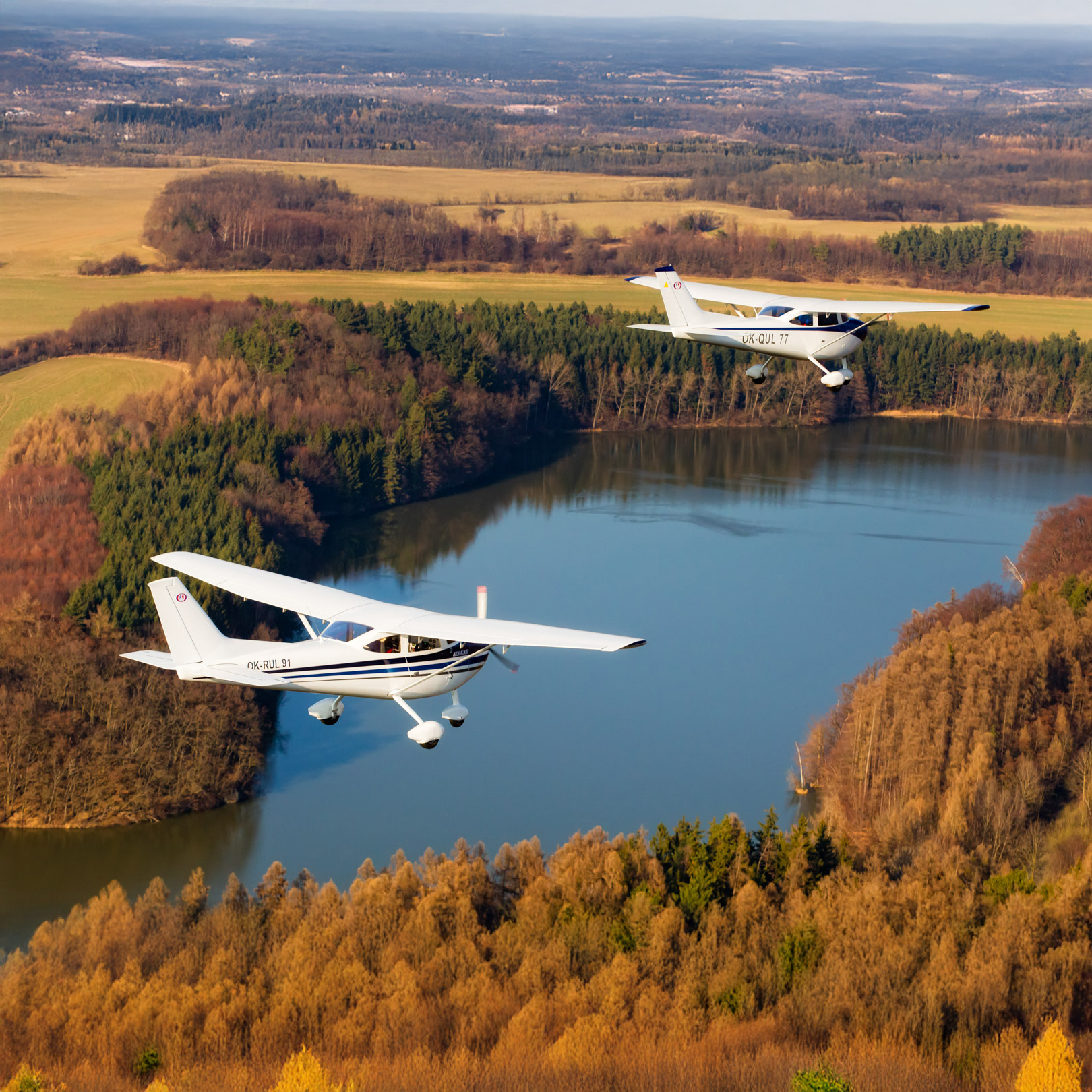 Zwei Leichtflugzeuge vom Typ Legend im Flug über eine herbstliche Waldlandschaft mit See, bei klarem Wetter.