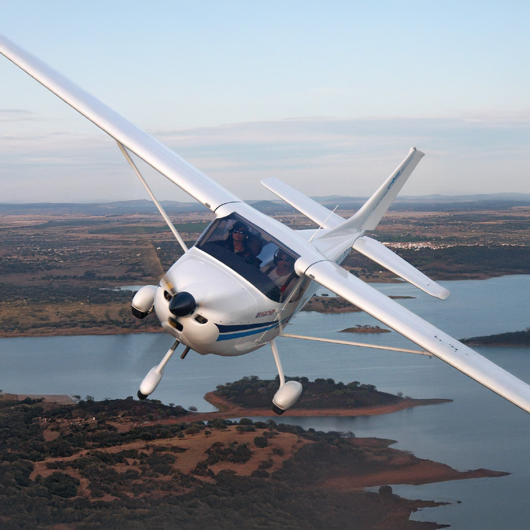 Weißes Leichtflugzeug mit zwei Personen im Cockpit im Flug über eine hügelige Landschaft mit Seen bei ruhigem Wetter.