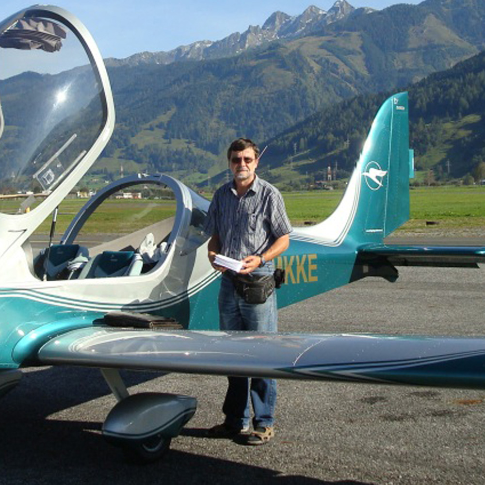Mann mit Sonnenbrille und Unterlagen in der Hand steht vor einem offenen Leichtflugzeug auf einem Flugplatz mit Bergkulisse im Hintergrund.