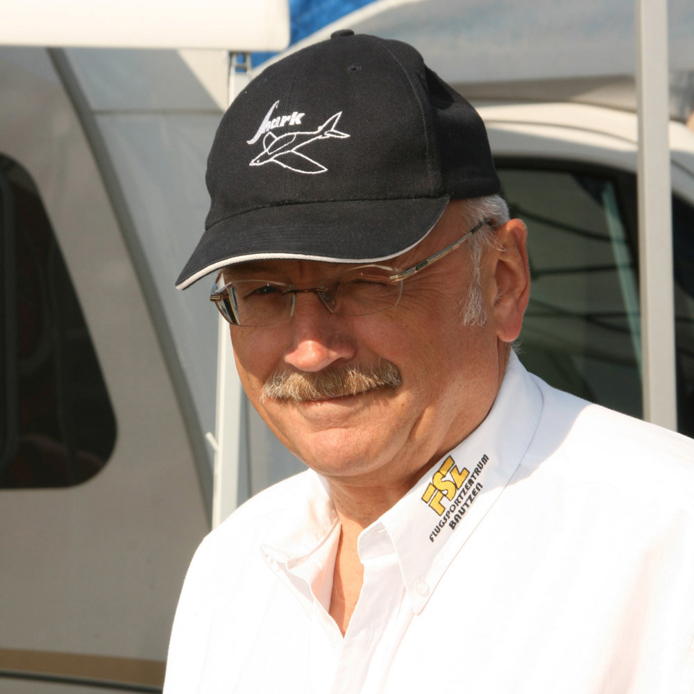 Mann mit Brille, weißem Hemd mit FSZ-Logo und dunkler Baseballkappe mit Flugzeugmotiv, im Freien vor einem Fahrzeug stehend.