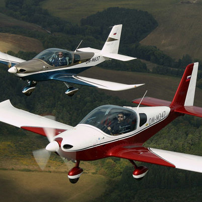Zwei einmotorige Leichtflugzeuge in Formation über einer hügeligen Landschaft, eines rot-weiß, das andere blau-weiß lackiert.